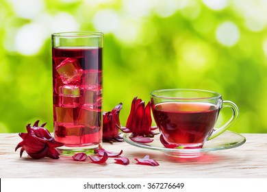 Cup of hot hibiscus tea (karkade, red sorrel, Agua de flor de Jamaica) and the same cold drink with ice cubes in glass on nature background. Magenta-color calyces (sepals) of roselle flowers on table.