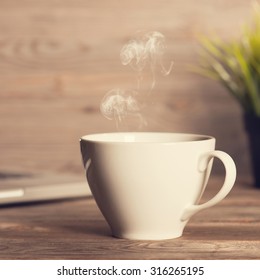 Cup Of Hot Steaming Coffee On Working Table, Laptop And Plant Pot At Background. Wooden Desk In Soft Focus With Noise Vintage Toned.