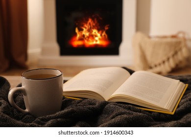 Cup of hot tea and book on knitted plaid near fireplace at home. Cozy atmosphere