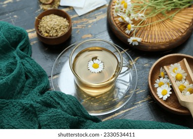 Cup of hot tea and bowl with dried chamomile on black wooden background