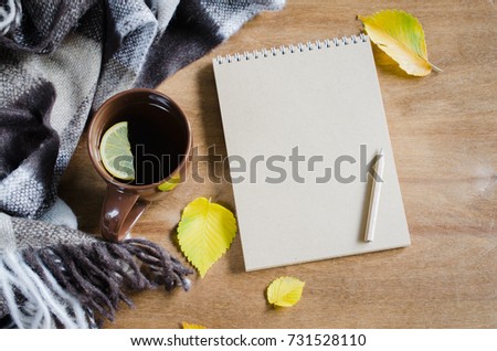 A cup of hot tea with lemon and empty notebook for sketch, plaid on a rustic table, yellow leaves. Cozy autumnal mood, warm autumn.