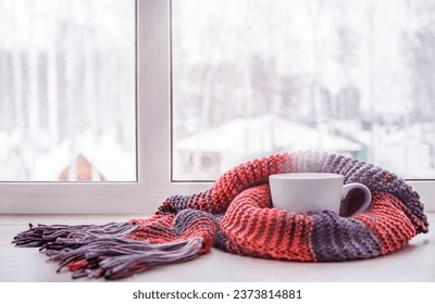 A cup of hot tea warmed in the knitted scarf is on the windowsill against the winter window