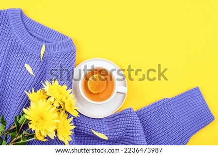 A cup of lemon tea, yellow chrysanthemum flowers on purple knitted sweater on yellow background. Top view, flat lay, copy space