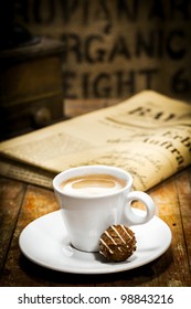 Cup Of Rich Milky Coffee With A Chocolate Bonbon On The Saucer And A Folded Newspaper On The Table Behind In A Coffee Break Concept