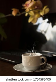 A Cup Of Steaming Coffee On The Working Table Near A Laptop