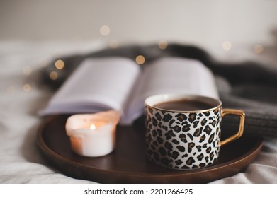 Cup Of Tasty Coffee On Wooden Tray With Open Paper Book And Burning Scented Candle In Bed Close Up. Cozy Home Hygge Atmosphere. Winter Season. 