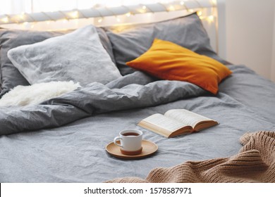 Cup Of Tea And Book Lie On The Bed With Gray Linen, Blanket, Pillows, Plaid And Led Garland Light. Cozy Winter Home Scene. Reading In Bed.