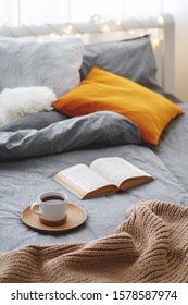 Cup Of Tea And Book Lie On The Bed With Gray Linen, Blanket, Pillows, Plaid And Led Garland Light. Cozy Winter Home Scene. Reading In Bed.