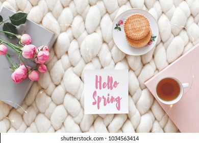 Cup Of Tea, Cookies, Pink Rose Flowers, Book And Card With Text Hello Spring On White Knitted Woolen Merino Chunky Blanket. Breakfast In Bed. Cozy Spring Homely Scene. Flat Lay, Top View. Home Decor.