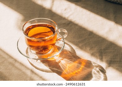 A cup of tea in glass cup and saucer illuminated by warm sunlight on a light table. Horizontal frame, copy space