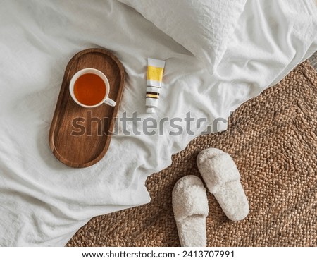 A cup of tea on a wooden tray, hand cream on the white sheets of the bed, slippers. Cozy house concept  