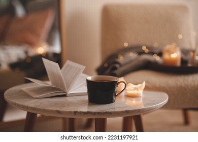 Cup of tea with paper open book and burning scented candles on marble table over cozy chair and glowing lights in bedroom closeup. Winter holiday season.  - Shutterstock ID 2231197921