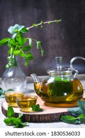 Cup and teapot of herbal tea with fresh mint leaves on wooden table. Dark background.