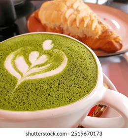 A cup of vibrant green matcha latte with intricate latte art sits in the foreground, accompanied by a flaky almond croissant on a plate in the background. - Powered by Shutterstock