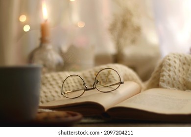 Cup of warm drink, chocolate chip cookies, open book and reading glasses. Candles, flowers and bokeh lights in the background. Hygge at home. Selective focus. - Powered by Shutterstock