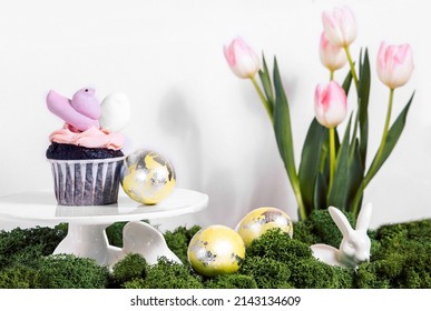 Cupcake With A Bird Marshmallow Peeps With An Egg On A Cake Stand In The Shape Of A Rabbit. Easter Greeting Card. Ceramic White Rabbit In Green Moss On A Background Of Pink Delicate Tulips