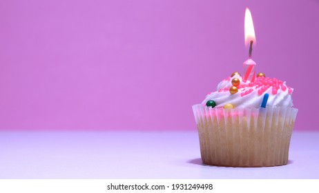 Cupcake With Burning Festive Candle At White Cream And Sugar Icing On Pink Background, Macro Shallow Depth Of Field View. Homemade Vanilla Cup Cake With Buttercream Frosting Close Up. 