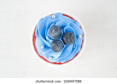 Cupcake Red Velvet With Blue Whipped Cream Decorated With Blueberry, Silver Confectionery Balls On White Wood Table. Picture For A Menu Or A Confectionery Catalog. Top View.