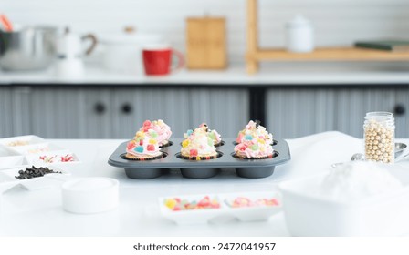 Cupcakes with pastel color butter cream, colorful jelly and chocolate flakes on baking tray in kitchen at home. Flour, sugar and decorating candy on table. Selective focus - Powered by Shutterstock