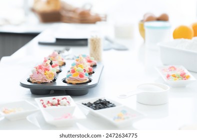 Cupcakes with pastel color butter cream, colorful jelly and chocolate flakes on baking tray in kitchen at home. Flour, sugar, eggs and decorating icing candy on table. Selective focus - Powered by Shutterstock