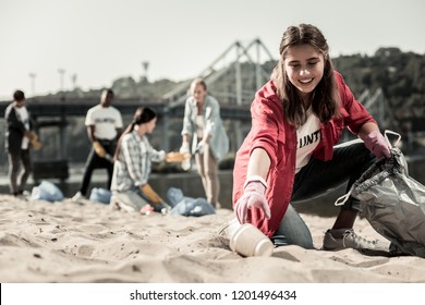 Cups in bag. Young dark-haired student wearing red anorak putting cups into garbage bag while volunteering on the beach