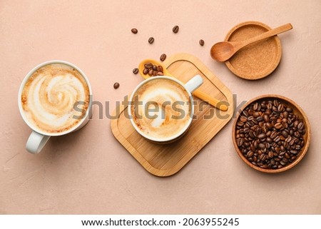 Cups of hot cappuccino and plate with coffee beans on color background