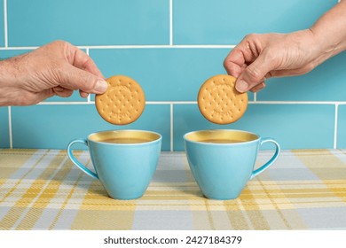 Cups of tea in attractive blue and yellow cups with hands dunking biscuits. Cups isolated against a blue tiled background and sitting on yellow check. Mugs of hot drink with a sweet treat.  - Powered by Shutterstock