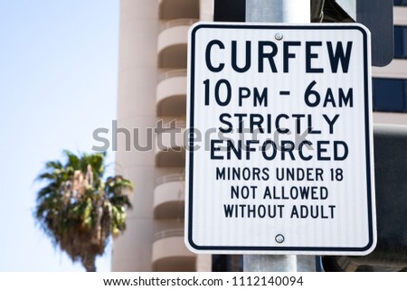 Curfew 10pm to 6am Strictly Enforced Minors Under 18 Not Allowed Without Adult sign, on a downtown city street, with palm tree in the background and space for text on left