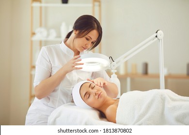 Curing skin problems. Cropped female cosmetologist looking at client's face through magnifying lamp examining her skin. Happy relaxed young woman getting professional facial treatment in spa salon - Shutterstock ID 1813870802