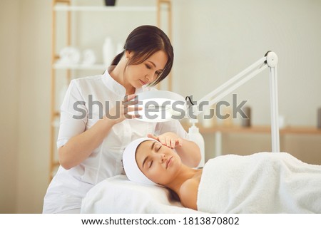 Curing skin problems. Cropped female cosmetologist looking at client's face through magnifying lamp examining her skin. Happy relaxed young woman getting professional facial treatment in spa salon