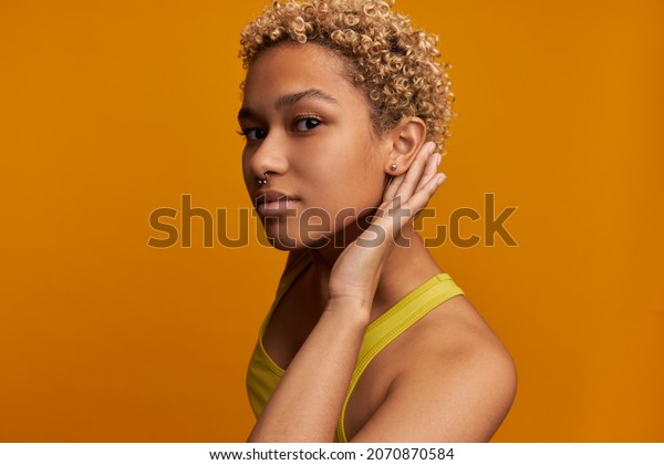 Curious African American lady in yellow summer\
clothes holding hand over ear trying to hear more, listening\
gossips, rumors, looking at camera with concentrated face\
expression. Body language\
concept