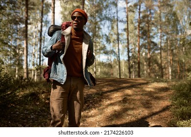Curious african american tourist guy wander among trees with backpack in hat and sunglasses, happy man enjoying time alone in green forest. Escape from hectic life to wildlife. Search for adventures - Shutterstock ID 2174226103