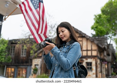 Curious Asian Woman Visitor Looking Into Distance With Camera In Hand While Photographing Street Scenes Of The Fairytale Town Carmel By The Sea Against America Flag Background