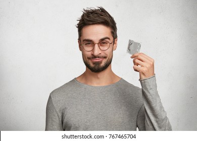 Curious bearded male holds new kind of condom, wonders about its quality, wears casual grey sweater, stands against white concrete wall. Handsome young man advertises product of contraception - Powered by Shutterstock