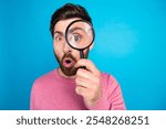 Curious bearded man exploring with magnifying glass against vibrant blue background, playful expression