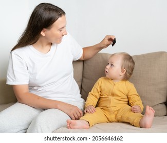 Curious Blond Child In Yellow Clothing Looking With Interest At Caucasian Light-haired Young Mother In Casual Grooming Baby Hair With Comb In Hand. Mother Care. Concept Of Parenthood