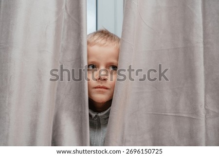 Curious boy peeks between gray curtains, half-body portrait capturing quiet, playful moment in a home interior