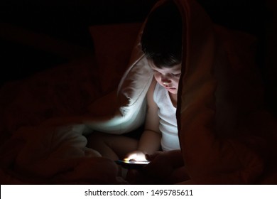 Curious Boy Sits In Bed Under White Blanket And Play Games On Smartphone In The Dark Room. The Child's Face Is Illuminated By A Bright Monitor. Toddler Holding Mobile In Hands. 