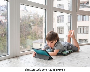 Curious Boy Watch Cartoons On Digital Tablet. Kid Lies On Floor And Uses Electronic Device. Indoor Leisure For Children While It's Raining Outside.