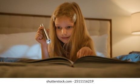 Curious Caucasian child little girl reading book instructive story textbook daughter lying bed learning home distant education studying homework kid holding flashlight evening night bedroom indoors - Powered by Shutterstock