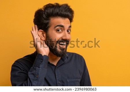 Curious Cheerful Indian Man Holding Hand Near Ear Trying To Overhear Something, Excited Young Eastern Guy Interested In Fresh Gossips, Standing Against Yellow Studio Background, Closeup Shot