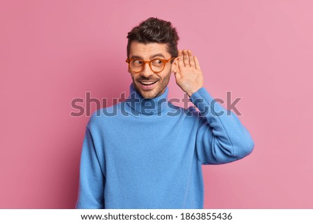 Curious cheerful unshaven man keeps hand near ear tries to overhear private conversation listens gossips dressed in casual blue poloneck isolated over pink background being interested in something