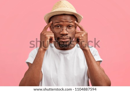 Curious dark skinned middle aged male keeps fore fingers on temples, wears straw hat, poses against pink background, tries to remember something important, has thoughtful expression, clever look
