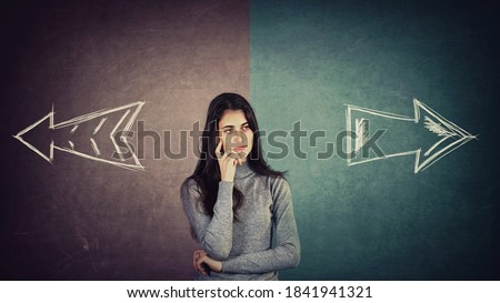 Curious and doubtful young woman, has to choose right or left side. Puzzled student girl in front of a split blackboard with arrows showing two different directions. Choice concept, difficult decision
