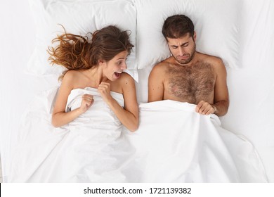 Curious excited woman looks at mans genital while lie in bed together. Displeased man looks under white blanket at penis, suffers from sexual dysfunction. Sex problems, marriage, relationship concept