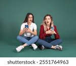 Curious friends, full body shocked surprised two young caucasian women curious friends sit floor look each others phone with open mouth. Green studio background, copy space.