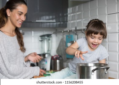 Curious Funny Kid Girl Playing With Pot With Mom In The Kitchen, Excited Active Child Helping Single Mother Preparing Food With Cookware, Family Mommy And Daughter Having Fun Cooking Together