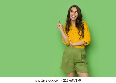 Curious and happy beautiful young woman in linen shirt and shorts looking to the side and talking. Three quarter length shot against green background. - Shutterstock ID 2167697179