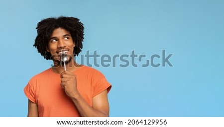 Curious and hungry young african american man with spoon in his mouth looking at copy space, dreaming about delicious healthy food, choosing something to eat, blue studio background, panorama