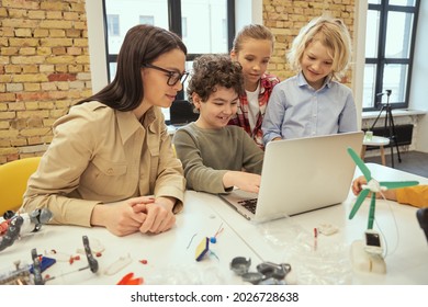 Curious kids using laptop, watching scientific robotics video, sitting at the table in a classroom together with young female teacher during STEM class